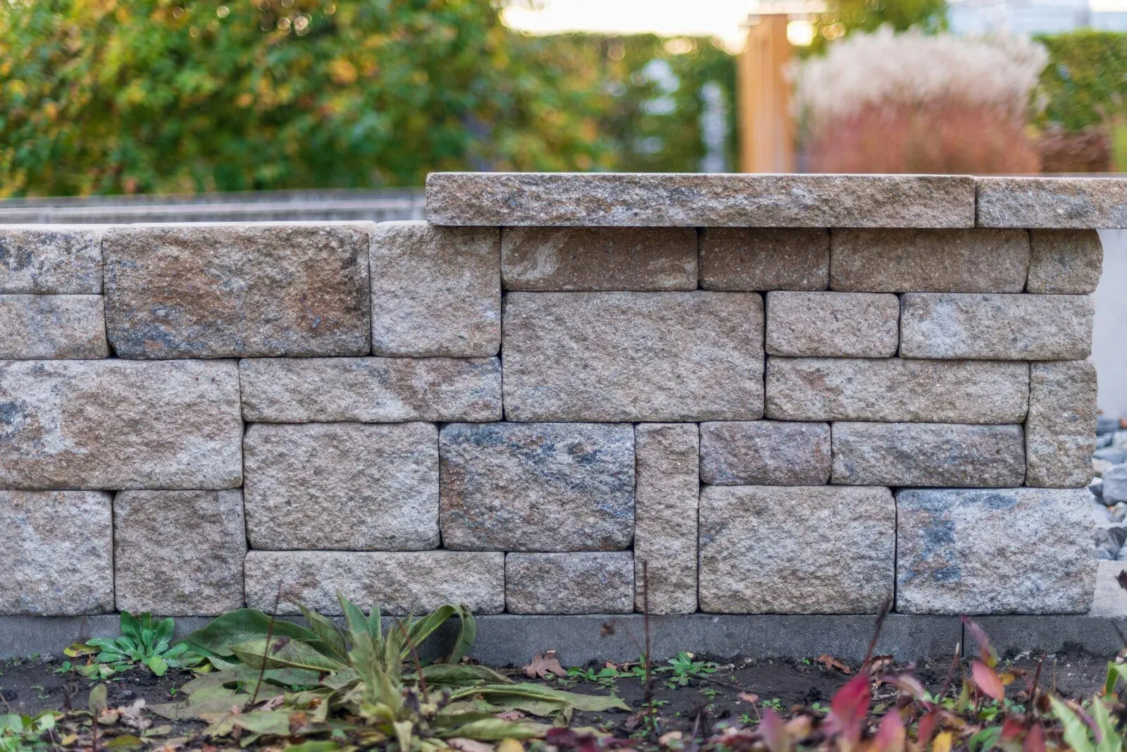 Segmental block retaining wall in tan and gray tones with a flat capstone course and plants along its base