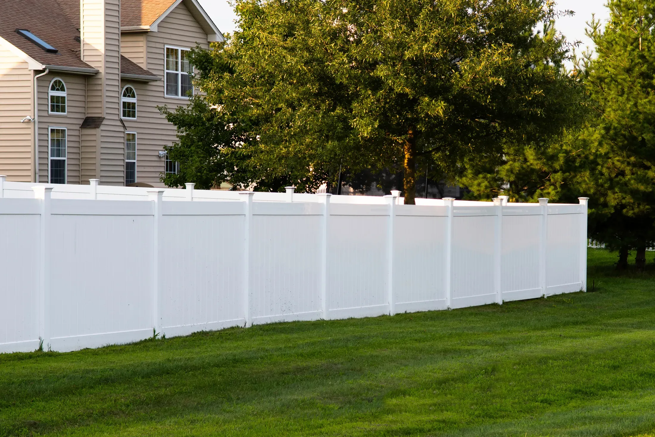 White vinyl privacy fence running along a residential lawn with houses and trees behind it